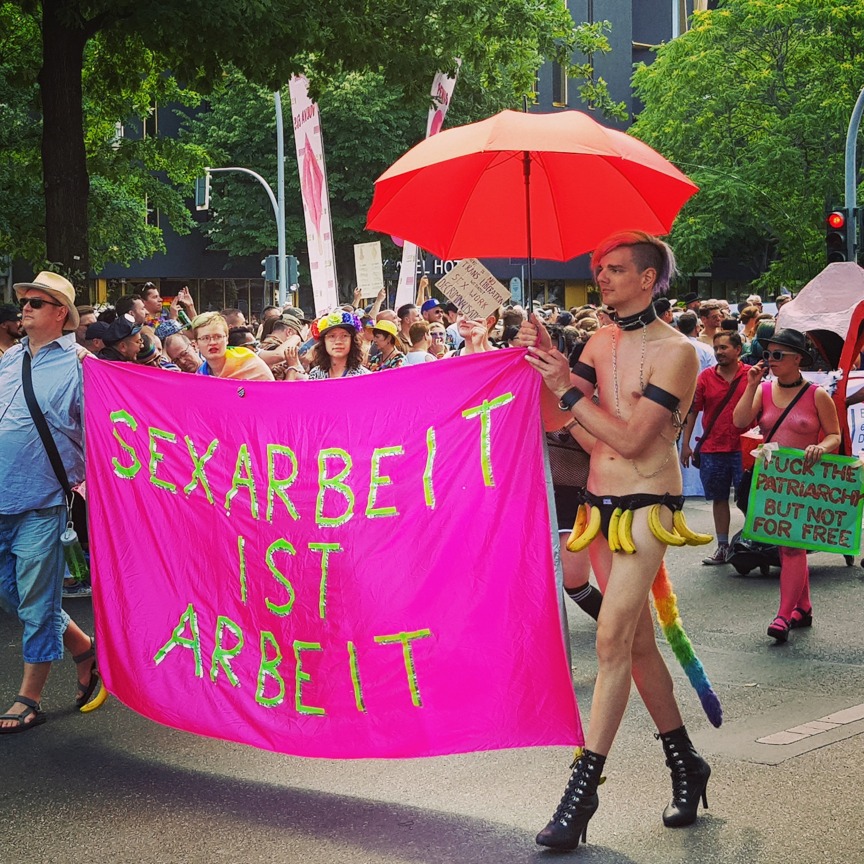 ein Transparent 'Sexarbeit ist Arbeit' auf dem Berliner CSD 2019 gehalten von einem Menschen in Jeans und Hemd - und einem in Jockstrap und High-Heels (ich). Im Hintergrund ein Plakat 'Fuck the patriarchy but not for free'